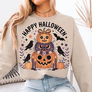 Puede incluir: Sudadera beige con un diseño de calabazas apiladas. El diseño incluye tres calabazas, un murciélago, cuervos y las palabras "Happy Halloween". Las calabazas son naranjas, moradas y decoradas con elementos florales.
