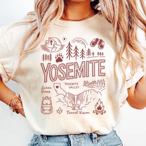 May include: Cream-colored t-shirt with a maroon graphic design featuring the word "YOSEMITE" and illustrations of a tent, trees, binoculars, a paw print, a waterfall, and a campfire. The shirt also includes the text "Sierra Views" and "Yosemite Valley".
