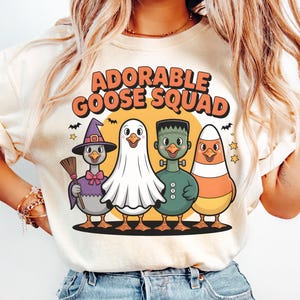 Könnte beinhalten: Cremefarbenes T-Shirt mit dem Text "ADORABLE GOOSE SQUAD" in Orange. Die Grafik zeigt vier Cartoon-Gänse in Halloween-Kostümen: eine Hexe, ein Geist, Frankenstein und ein Zuckermais.