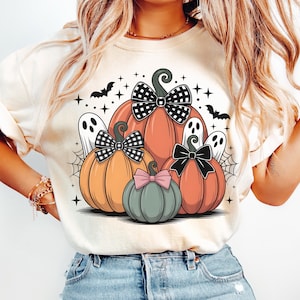 May include: Cream-colored t-shirt featuring a Halloween-themed graphic. The design includes orange, yellow, and gray pumpkins with bows, ghosts, bats, and spiderwebs. The pumpkins have black and white checkered, black, and pink bows.