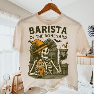 Peut inclure: T-shirt crème avec un motif d'un squelette barista portant un chapeau de sorcière, tenant deux boissons au café. Le texte "BARISTA OF THE BONEYARD" est imprimé au-dessus du motif. Un motif de chauve-souris est également inclus.