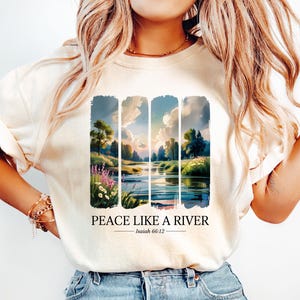 May include: Cream-coloured t-shirt with a scenic landscape design depicting a river, trees, and sky. The text "PEACE LIKE A RIVER" is printed below the artwork, along with the biblical reference Isaiah 66:12.