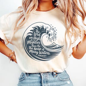 Könnte beinhalten: Cremefarbenes T-Shirt mit einem marineblauen Wellenmuster und dem Text "The Lord on High is Mightier than the Noise of Many Waters. Psalm 93:4." Das Shirt hat kurze Ärmel und eine lockere Passform, geeignet für den Alltag.