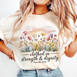 Puede incluir: Camiseta color crema con un diseño floral en acuarela. El diseño incluye flores rojas, moradas y amarillas. El texto "clothed in strength & dignity" está impreso debajo de las flores, con "Proverbios 31:25" debajo.