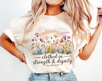 Może przedstawiać: Kremowa koszulka z akwarelowym wzorem kwiatowym. Wzór zawiera czerwone, fioletowe i żółte kwiaty. Tekst "clothed in strength & dignity" jest wydrukowany pod kwiatami, z "Proverbs 31:25" pod spodem.