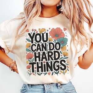 May include: Cream-colored t-shirt with the text "YOU CAN DO HARD THINGS" in black, surrounded by colorful floral and heart graphics. The design includes pink, yellow, and blue flowers, and green leaves. A casual, inspirational tee.
