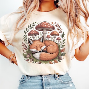 May include: Cream-colored t-shirt featuring a whimsical illustration of a sleeping fox nestled among mushrooms, leaves, and acorns. The fox has orange fur and is curled up. The mushrooms have brown caps with white spots. The design is surrounded by green foliage.