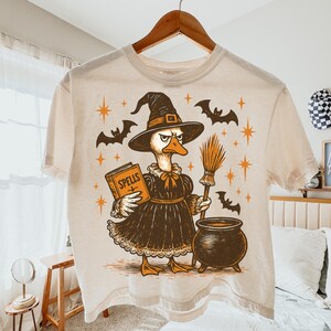 Könnte beinhalten: Cremefarbenes T-Shirt mit einer Cartoon-Ente, die als Hexe verkleidet ist und ein Zauberbuch und einen Besen hält. Das Design umfasst Fledermäuse, Sterne und einen Kessel, ideal für Halloween.