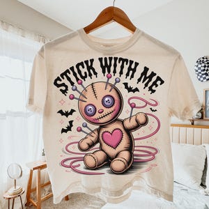 May include: Cream-colored t-shirt featuring a cartoon voodoo doll with pins, a heart, and the text "STICK WITH ME." The design includes bats and pink accents. The shirt is hanging on a wooden hanger.