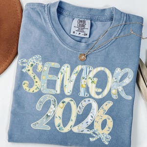 Puede incluir: Una camiseta Comfort Colors azul claro con las palabras "SENIOR 2026" en un diseño floral y a rayas. Un collar dorado con un colgante está drapeado sobre la camisa. Un sombrero marrón y una planta seca están en el fondo.