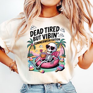 Pode incluir: Camiseta creme com um esqueleto usando óculos de sol em forma de coração, relaxando em uma boia inflável rosa. O gráfico inclui palmeiras, fantasmas e o texto "DEAD TIRED BUT VIBIN' SUMMERWEEN EDITION."
