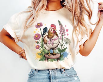 May include: Cream-colored t-shirt featuring a detailed illustration of a hen standing on a wooden stool amidst colorful flowers and greenery. The hen wears a pink and white checkered scarf. Watercolor paint splatters add a whimsical touch to the design.