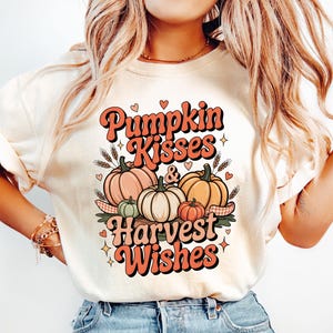 May include: Cream-colored t-shirt with the text "Pumpkin Kisses & Harvest Wishes" in orange and black. The graphic features pumpkins in various sizes and colors, with wheat stalks and hearts.