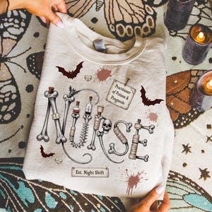 May include: A light beige sweatshirt with a spooky design. The word "NURSE" is spelled out with bones and medical equipment. Includes bats, blood splatters, and the text "Practitioner of Potions & Prognosis" and "Est. Night Shift".