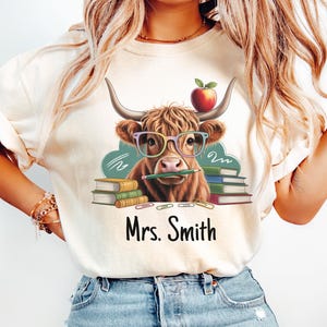 Könnte beinhalten: Cremefarbenes T-Shirt mit einer Cartoon-Highland-Kuh, die eine Brille trägt und einen Bleistift im Mund hält. Ein Apfel sitzt auf ihrem Kopf. Der Text "Mrs. Smith" ist unter dem Kuhbild aufgedruckt, mit Bücherstapeln auf beiden Seiten.