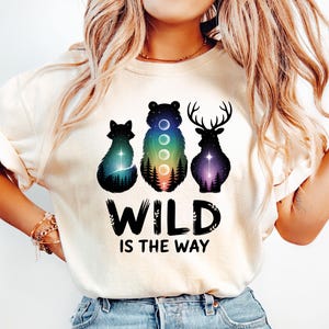 Może przedstawiać: Kremowa koszulka z grafiką lisa, niedźwiedzia i jelenia z niebiańskimi wzorami. Tekst "WILD IS THE WAY" jest wydrukowany na czarno pod ilustracjami zwierząt. Koszulka ma podwinięte rękawy.