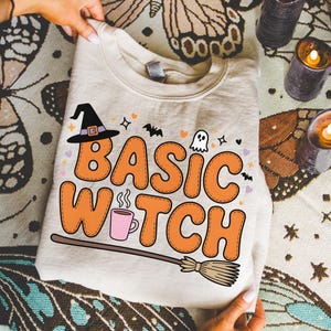 Può includere: Una felpa color crema con la scritta "BASIC WITCH" in arancione, bordata di marrone. Il design include un cappello da strega, una tazza di caffè, una scopa e altre grafiche a tema Halloween. La felpa è esposta su una superficie a fantasia.