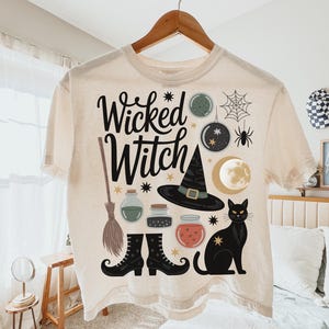 May include: Cream-colored t-shirt with the words "Wicked Witch" in black script. The shirt features Halloween-themed illustrations, including a black cat, witch's hat, broom, and potion bottles. A fun and festive design.