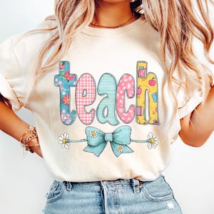 Op de afbeelding: Crèmekleurig T-shirt met het woord "teach" in kleurrijke, bedrukte letters. De letters hebben bloemen-, ruit- en stippenpatronen. Onder het woord zit een lichtblauwe strik met madeliefjes.