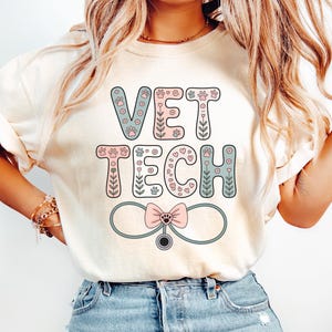 May include: Cream-colored t-shirt with the words "VET TECH" in pastel blue and pink bubble letters. The design includes paw prints, flowers, and a stethoscope with a pink bow. The shirt is a casual style.