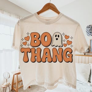 May include: Cream-colored t-shirt with the words "BOO THANG" in orange, outlined in black, and a ghost graphic. The design includes orange hearts and star shapes, perfect for Halloween.