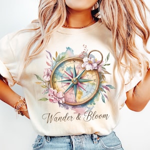 May include: A white t-shirt with a watercolor compass design and floral accents. The compass has a gold frame and blue, green, and pink accents. The text "Wander & Bloom" is printed below the compass.