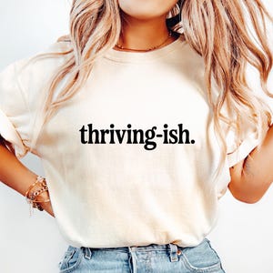 May include: Cream-colored t-shirt with the phrase "thriving-ish" printed in black. The shirt has rolled-up sleeves and is paired with blue jeans. The overall aesthetic is casual and comfortable.