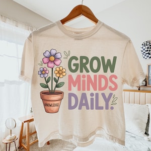 May include: Cream-colored t-shirt with a floral design and the words "GROW MINDS DAILY." The graphic features a potted plant with colorful flowers. The pot has the word "KNOWLEDGE" on it. The shirt is hanging on a wooden hanger.