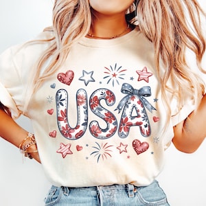 Puede incluir: Una camiseta blanca con un diseño gráfico de la palabra "USA" en letras florales rojas, blancas y azules. Las letras están rodeadas de corazones, estrellas y fuegos artificiales.