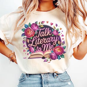 May include: Cream-colored t-shirt featuring a floral design with the text "Talk Literary to Me." The design includes purple and pink roses, leaves, and hearts surrounding an open book. The shirt is made of a soft material.