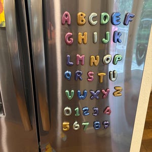 May include: A set of colorful, metallic-finish magnetic letters and numbers arranged in rows on a stainless steel refrigerator surface. The collection includes the full alphabet from A to Z and digits from 0 to 9, each featuring a rounded, inflated shape.