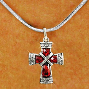 May include: A silver-toned pendant necklace featuring a cross design. The cross is adorned with red gemstones and clear crystals, suspended from a silver chain. The cross has a detailed, ornate design.