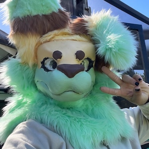 Może przedstawiać: Furry fursuit głowa z jasnozielonym, brązowym i żółtym schematem kolorów. Twarz jest jasnozielona z brązowymi akcentami, a uszy to mieszanka zieleni, brązu i żółci. Fursuit ma miękką, puszystą teksturę.