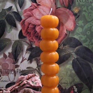 May include: A tall, orange candle with a stacked ball design, resting in a clear glass holder. The candle is set against a backdrop of floral wallpaper and a string of pearls.