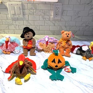May include: A collection of plush toys, including turkeys, a bear, and a pumpkin. The turkeys have colorful feathers in shades of pink, blue, and brown. The bear wears a hat and holds a small item. The pumpkin has a jack-o'-lantern face.