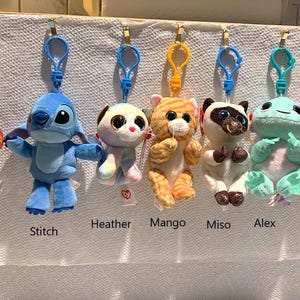 May include: A collection of plush toy keychains featuring various animal characters, including a pug, Stitch, a cat, and a raccoon. Each plush has a colorful clip for easy attachment. The names of the characters are displayed below each toy.