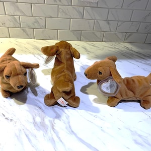 May include: Three brown dachshund stuffed animals with floppy ears and tags. The plush dogs are in various poses, showcasing their long bodies and short legs. The toys are on a white and gray surface.