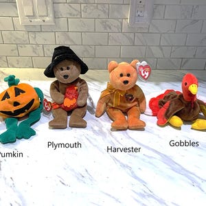 May include: Four Beanie Baby plush toys: a pumpkin, a bear wearing a witch hat, a bear with a bow, and a turkey. The pumpkin is orange and green. The bear toys are brown and orange. The turkey is brown, red, and yellow.