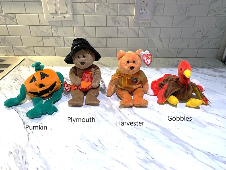 May include: Four Beanie Baby plush toys: a pumpkin, a bear wearing a witch hat, a bear with a bow, and a turkey. The pumpkin is orange and green. The bear toys are brown and orange. The turkey is brown, red, and yellow.