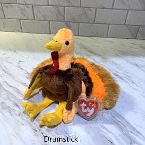 May include: A plush turkey toy with orange, brown, and yellow coloring. The turkey has a red wattle and a yellow beak. The toy has a tag that reads "TY Beanie Babies" and the word "Drumstick" is printed below the toy.
