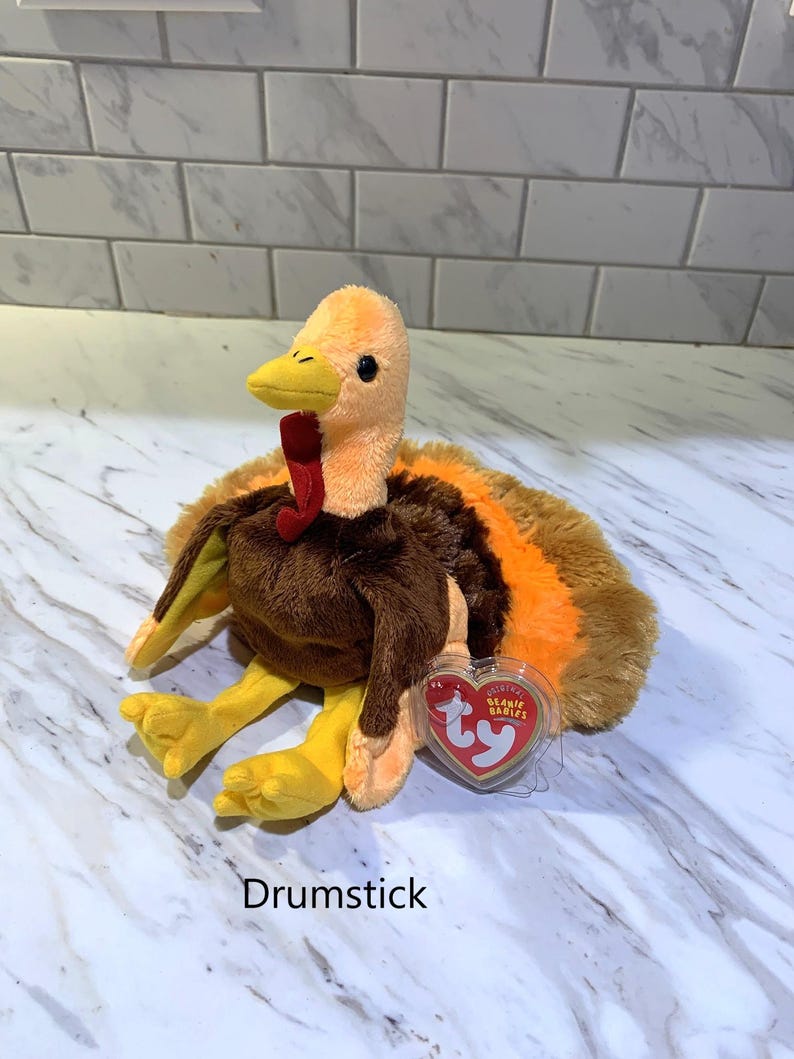 May include: A plush turkey toy with orange, brown, and yellow coloring. The turkey has a red wattle and a yellow beak. The toy has a tag that reads "TY Beanie Babies" and the word "Drumstick" is printed below the toy.
