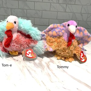 May include: Two plush turkey toys, "Tom-e" and "Tommy", are displayed. Tom-e has a pink and red body with a teal feather trim. Tommy has a brown body with purple and orange patterned feathers. Both have red snoods and orange beaks.