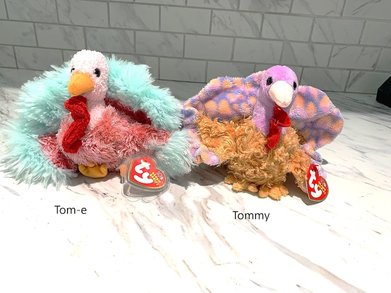 May include: Two plush turkey toys, "Tom-e" and "Tommy", are displayed. Tom-e has a pink and red body with a teal feather trim. Tommy has a brown body with purple and orange patterned feathers. Both have red snoods and orange beaks.