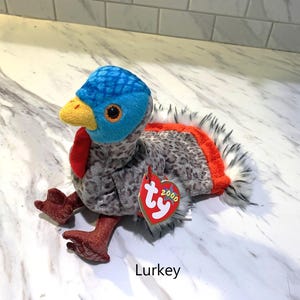 May include: A plush turkey toy named "Lurkey" with a blue head, yellow beak, and red wattle. The body is gray with a speckled pattern, and the tail feathers are white and black with an orange accent. A red heart-shaped tag is attached.