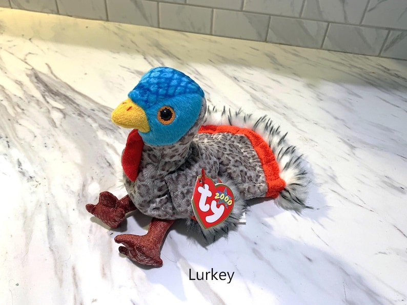 May include: A plush turkey toy named "Lurkey" with a blue head, yellow beak, and red wattle. The body is gray with a speckled pattern, and the tail feathers are white and black with an orange accent. A red heart-shaped tag is attached.