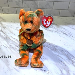 May include: A plush bear toy with orange and green striped fur, holding a leaf and acorn. The bear has a black nose and a red heart-shaped tag. The word "Leaves" is printed on the bottom left.
