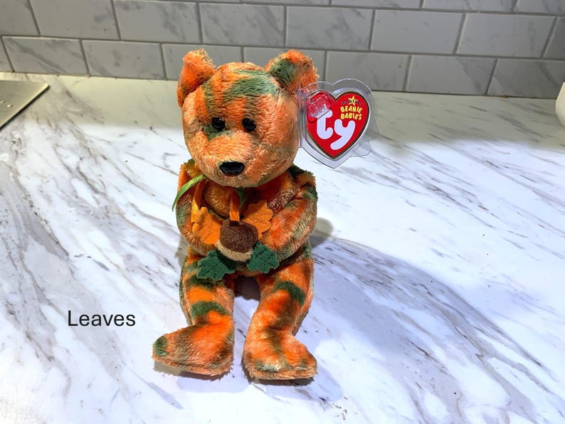 May include: A plush bear toy with orange and green striped fur, holding a leaf and acorn. The bear has a black nose and a red heart-shaped tag. The word "Leaves" is printed on the bottom left.