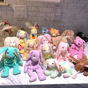 May include: A collection of colorful plush bunny toys in various sizes and colors, including pink, blue, yellow, and purple. Some bunnies have long ears and ribbons. The toys are displayed on a white countertop.