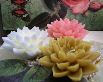 May include: Three decorative wax candles shaped like flowers. The candles are white, pink, and yellow. The candles are arranged on a surface with a pearl necklace.