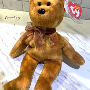 May include: A brown Ty Beanie Baby bear plush toy with a brown ribbon and the word "Thanksgiving" embroidered on its chest. The bear has a black nose and a red heart-shaped tag.